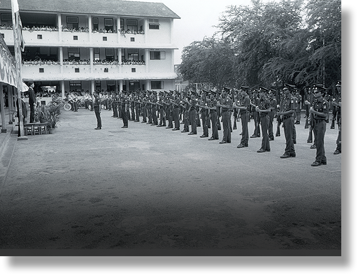 Black and white photo of uniformed soldiers standing in formation with rifles outside a multi-story building with spectators.