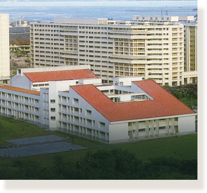 Aerial view of a white building with an orange-red roof, surrounded by tall multi-story buildings near a waterfront.
