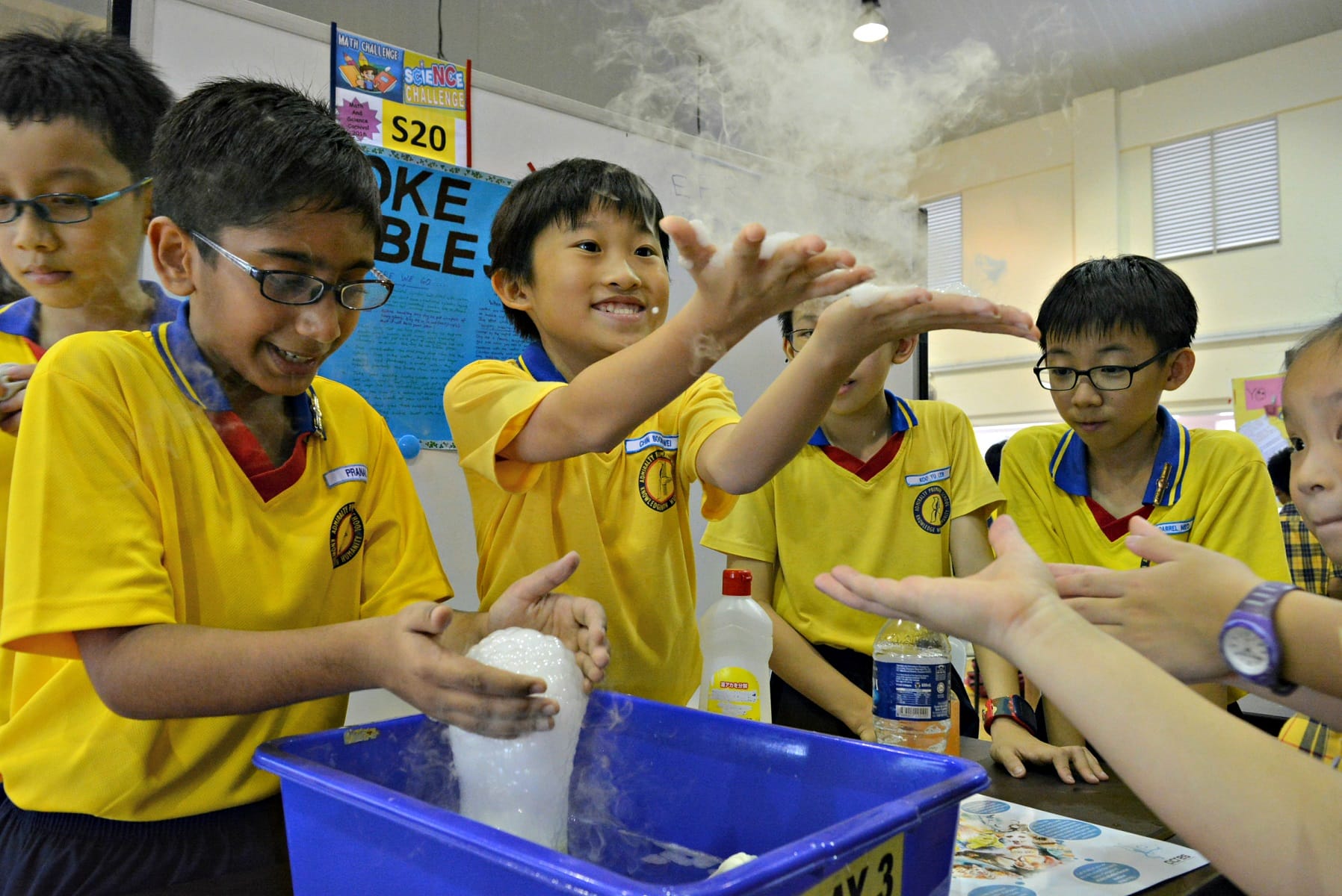 Children in yellow uniforms conduct a science experiment with dry ice producing smoke over a blue plastic tub.