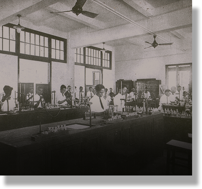 Vintage black and white photo of many people working at lab benches with equipment in a room with ceiling fans.