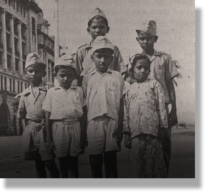 Black and white photo of six children in uniforms and caps posing together on a street with buildings behind them.