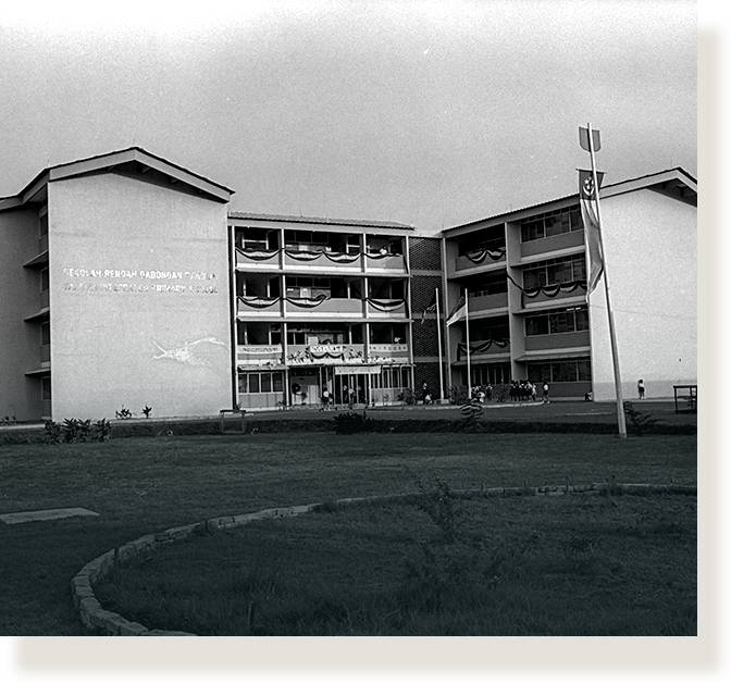 Black and white photo of a multi-story school building with a flagpole and open lawn in front.