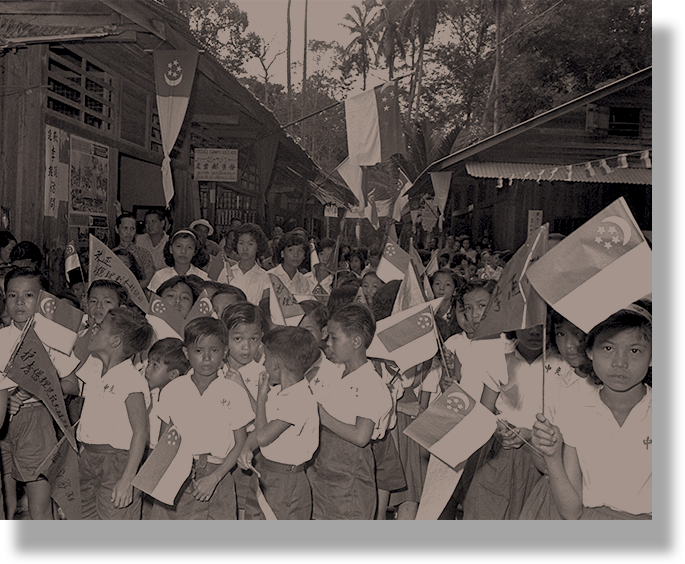Vintage sepia photo of children in uniforms marching through a street waving Singapore flags and banners.