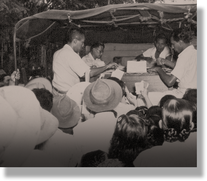 A vintage black and white photo of several people gathered around a table under a canopy, exchanging papers.