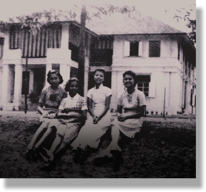 Four young women sit on grass in front of a two-story colonial-style building in a vintage black and white photo.
