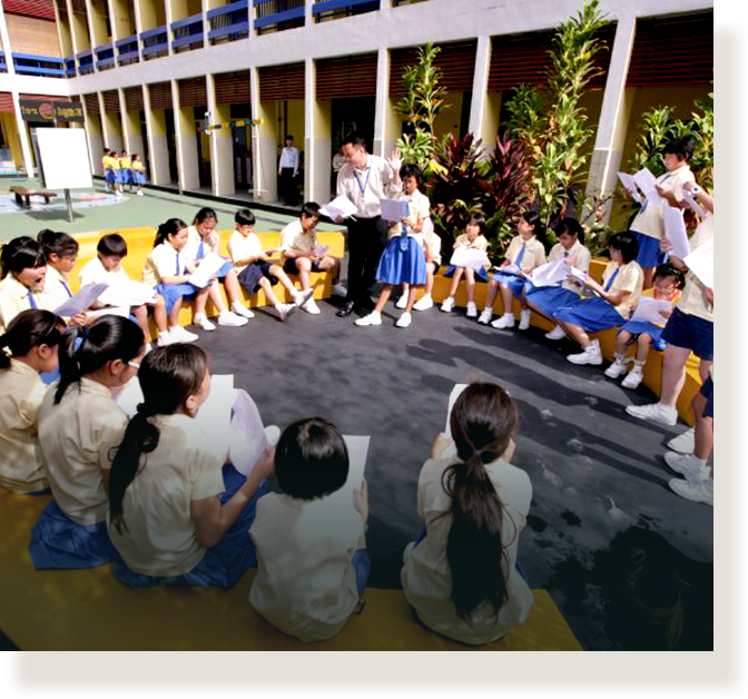 Students in uniforms sit in a circle outdoors while a teacher stands in the center holding papers.