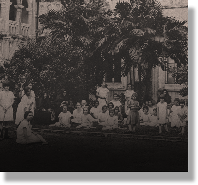 Old black and white photo of a large group of girls and women gathered outdoors near a building with palm trees.