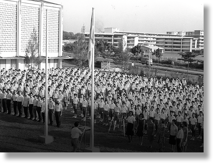 Black and white photo of hundreds of uniformed students assembled in rows on a field during a flag-raising ceremony.