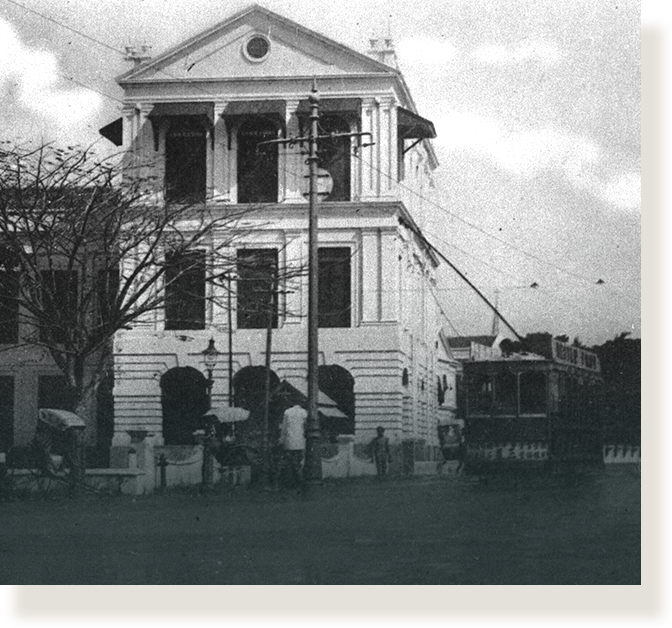 Black and white photo of a two-story colonial building with columns, a tram, and pedestrians outside.