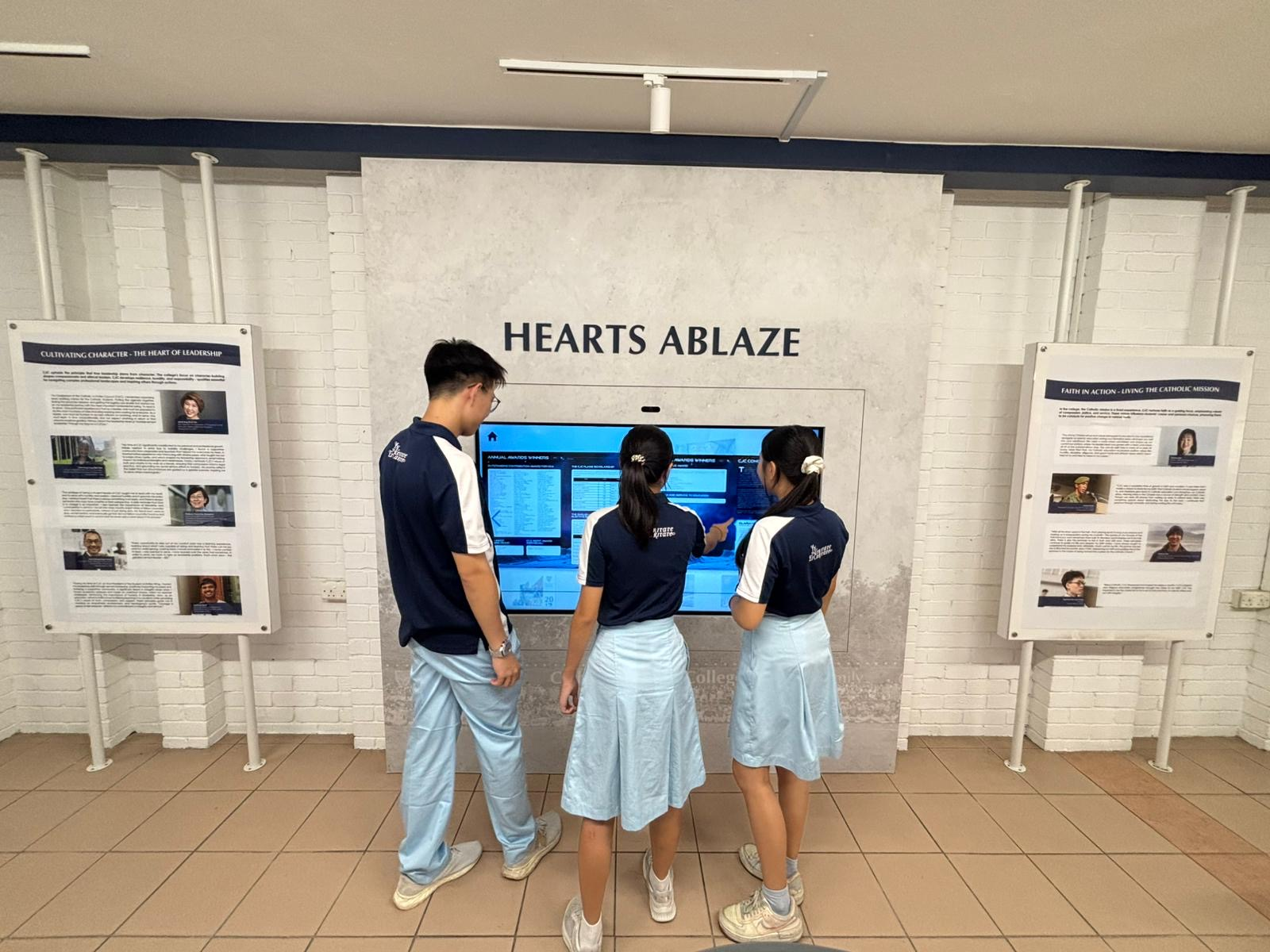 Three Catholic Junior College students in school uniform interact with the 'Hearts Ablaze' touchscreen kiosk, with one student pointing at the screen which displays college annuals content.