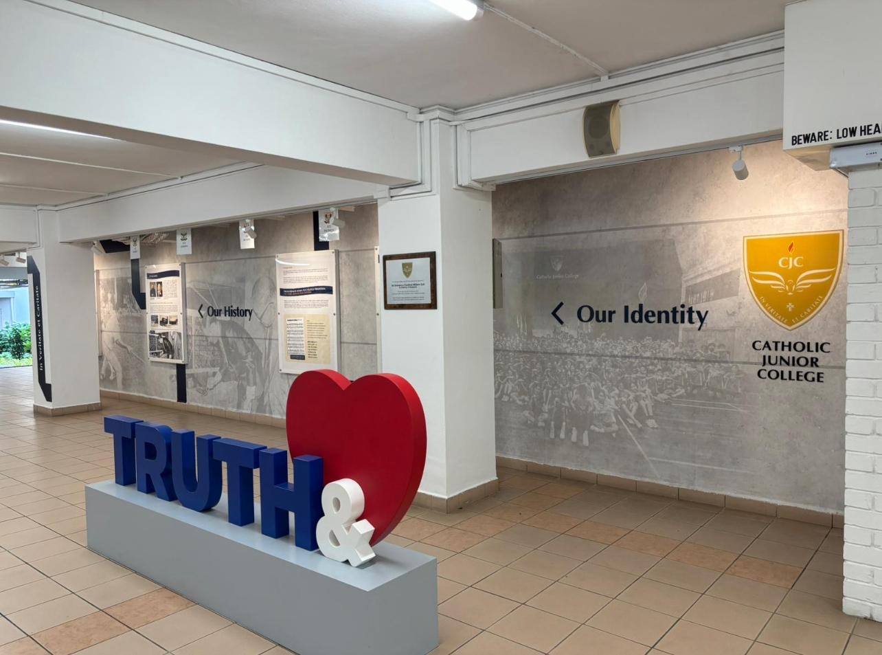 A corridor at Catholic Junior College featuring a large 3D sculpture of the words 'TRUTH &' in bold blue letters with a red heart symbol, placed on a light blue platform.