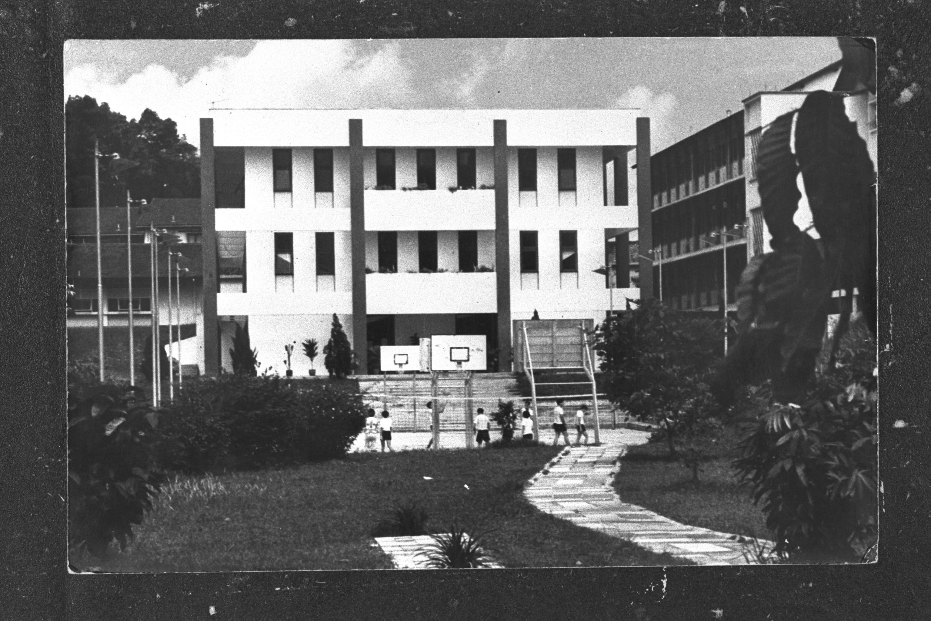 Black and white photo of a school compound with children near basketball hoops, a multi-story building, and a pathway.