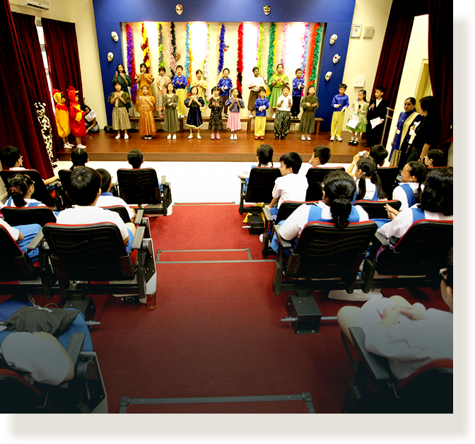 Children in costumes perform on a colorful stage while seated students in uniforms watch from an auditorium.