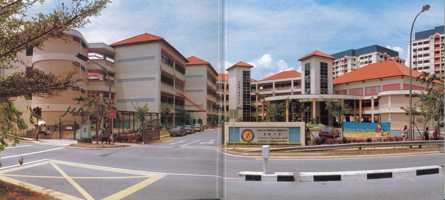 Admiralty Primary School building with red roofs, a sign at the entrance, and a road in front under blue sky.