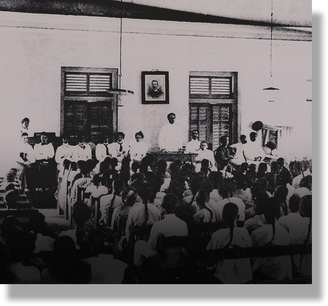 Black and white vintage photo of a large group gathered in a hall, with a speaker at a table and a framed portrait on the wall.
