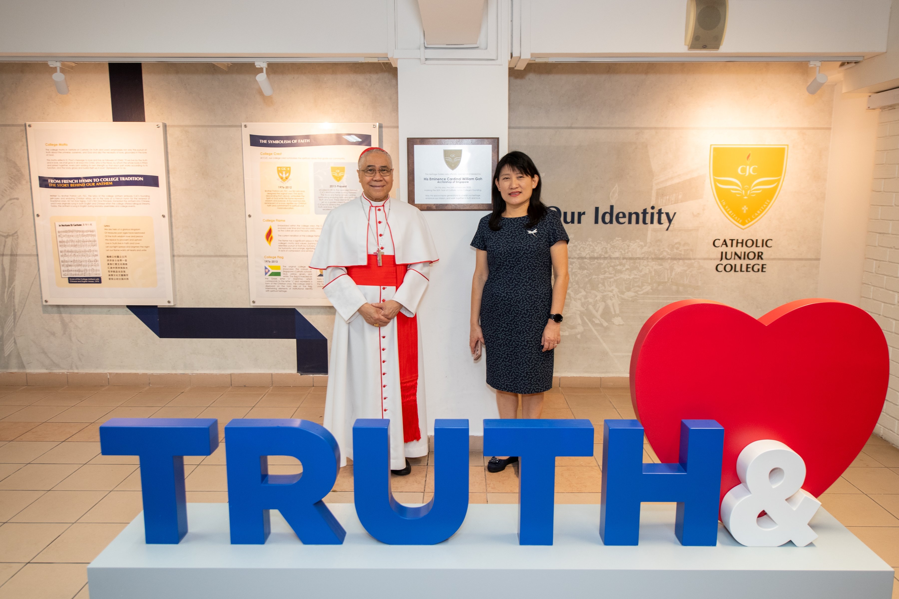 A Catholic cardinal in red and white vestments stands alongside a woman in a dark blue dress for a photo at the Catholic Junior College Heritage Gallery.