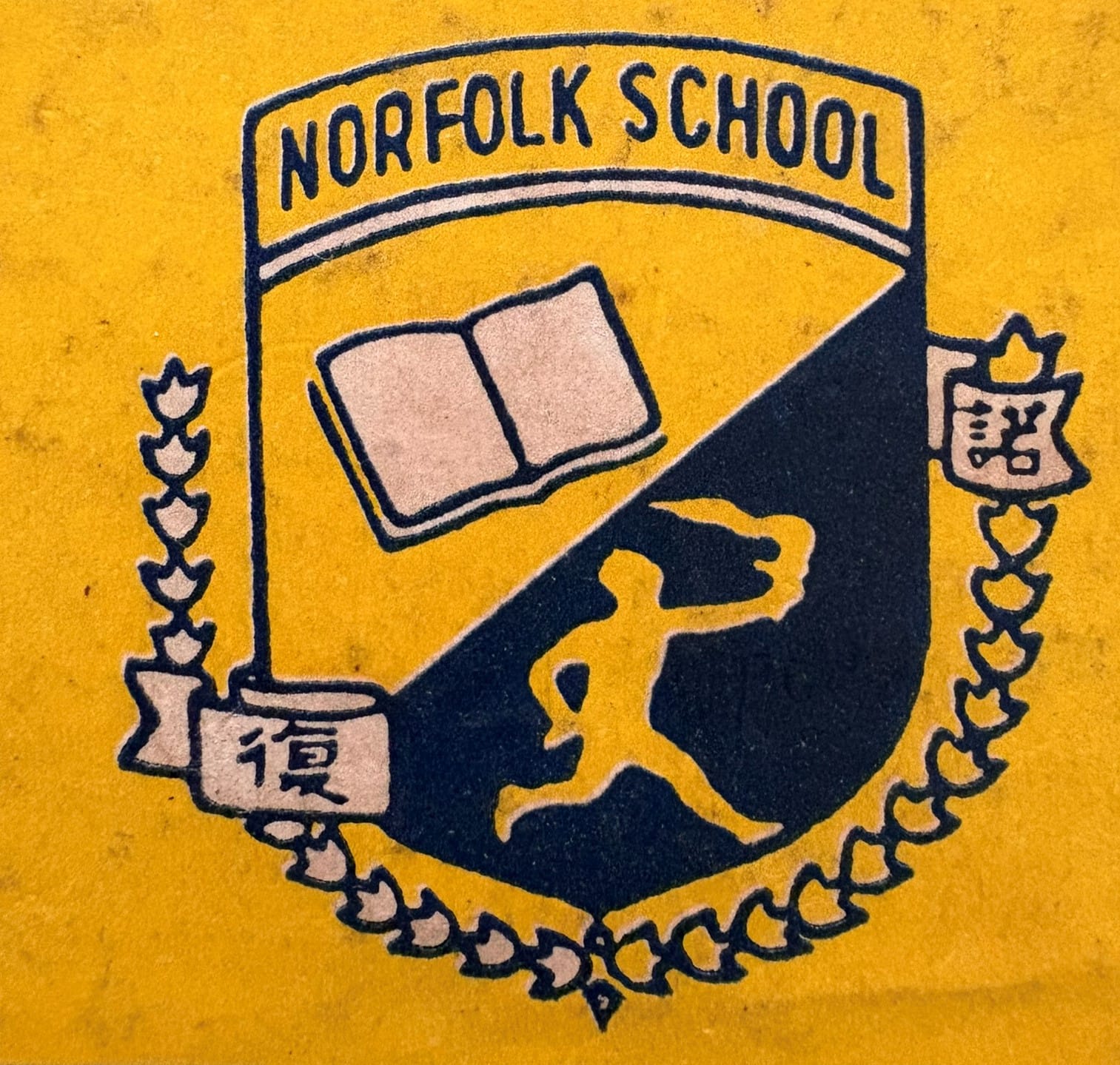 Norfolk School crest in navy and yellow showing a shield with an open book, a lion, laurel branches, and Chinese characters.