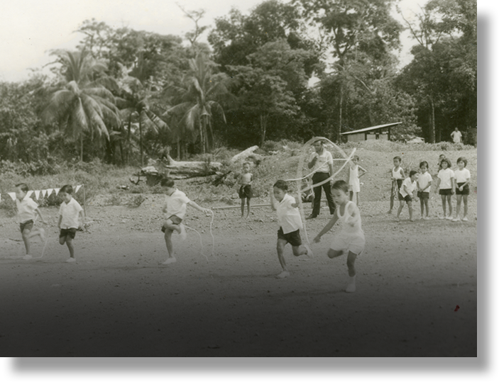 Black and white photo of children jump roping and playing outdoors on dirt ground with tropical trees behind them.