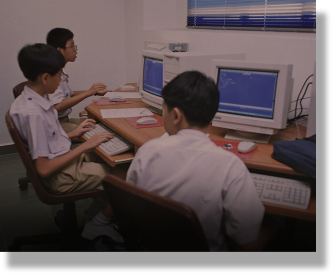 Three young students in white shirts sit at desks using older desktop computers with CRT monitors.