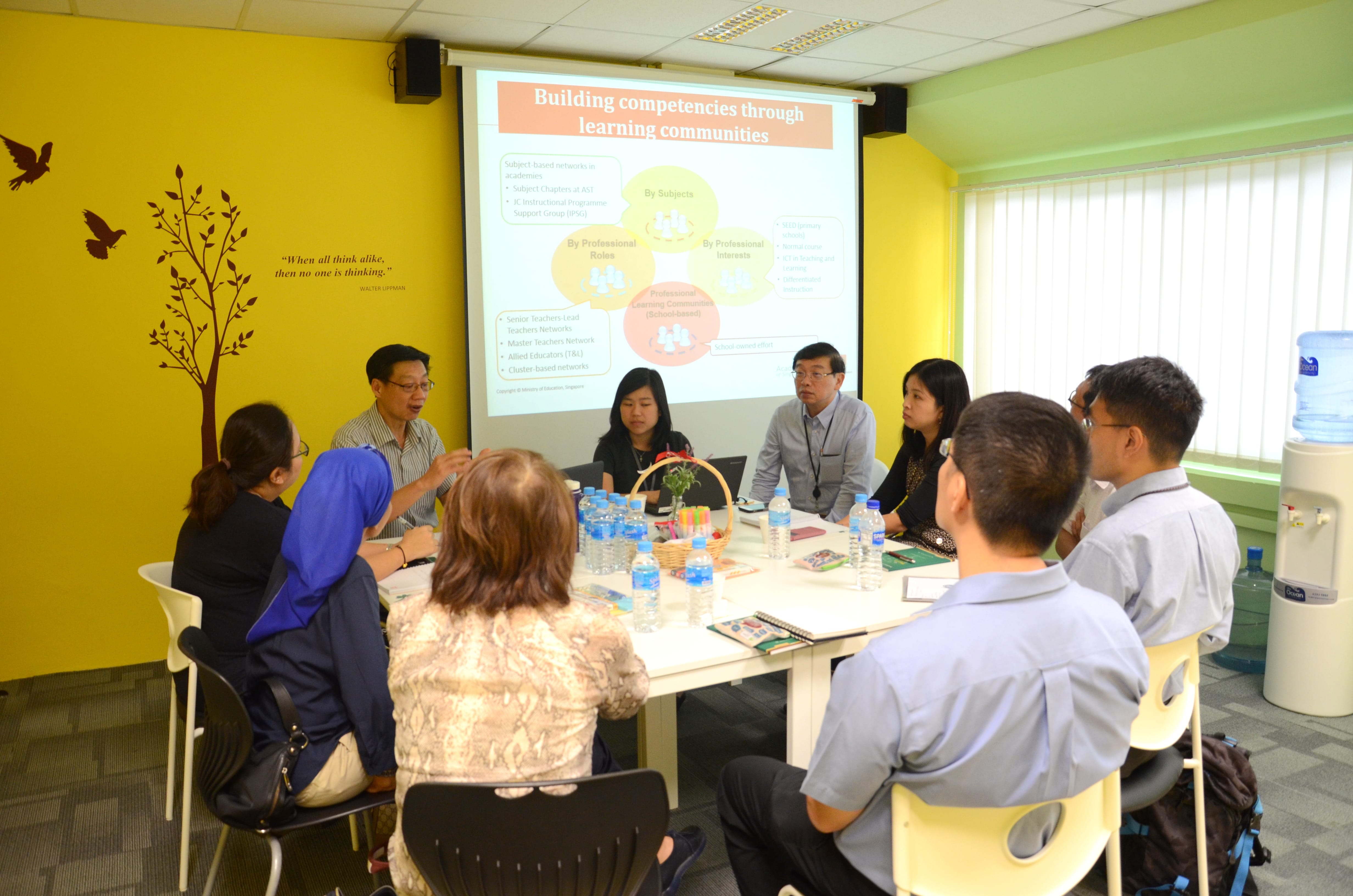 Nine people seated around a table in a meeting room, with a presentation slide projected on the wall.