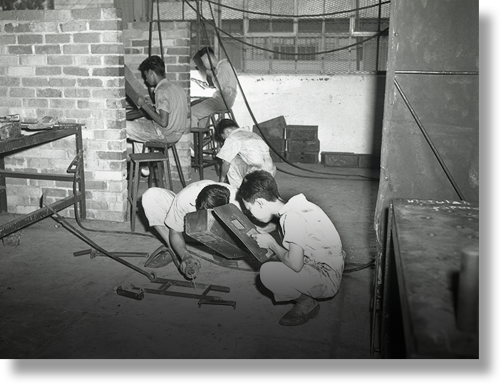 Black and white photo of young men working with tools and metal parts in a brick workshop.