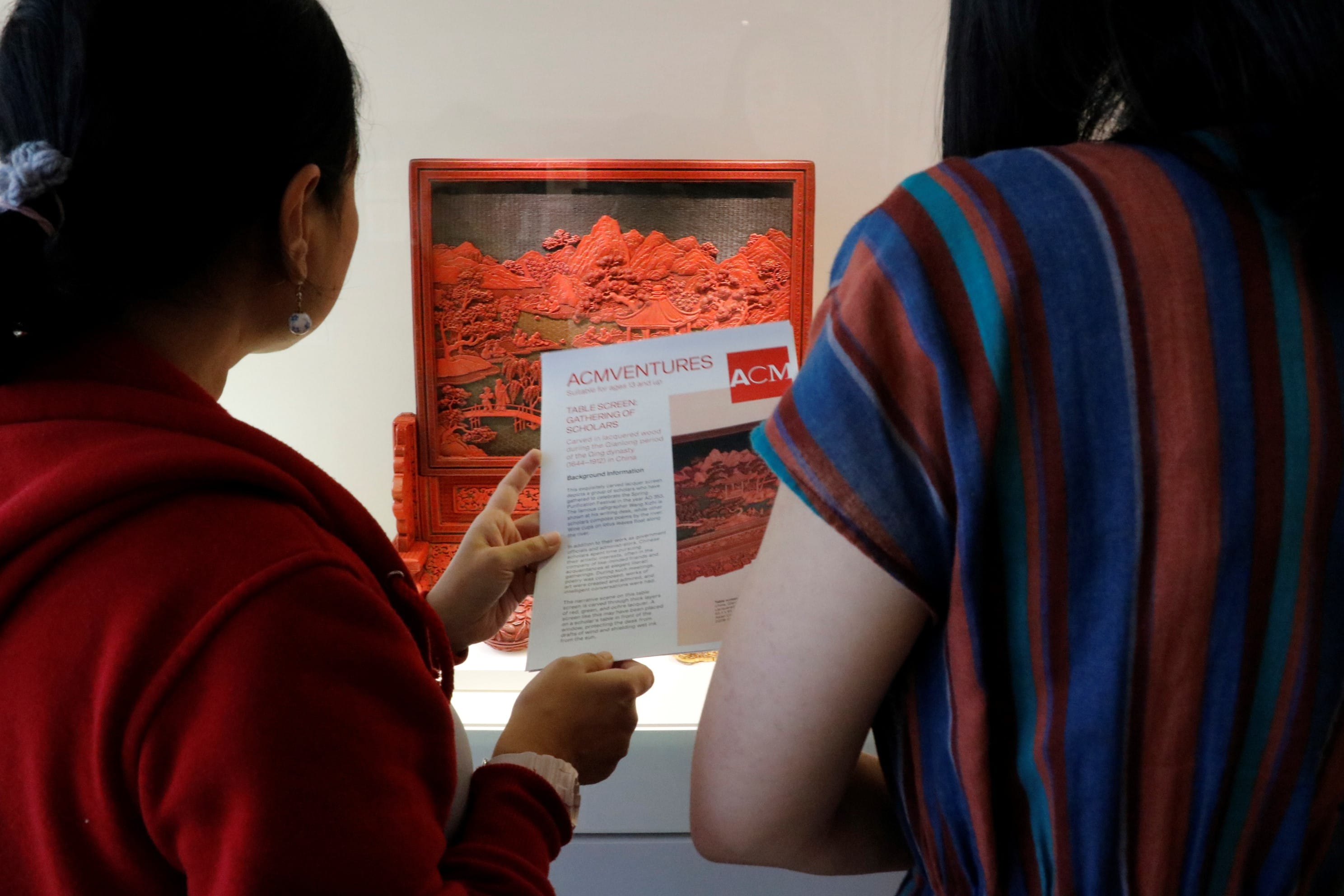 Two people read an ACMventures pamphlet in front of a red carved lacquer table screen on display.