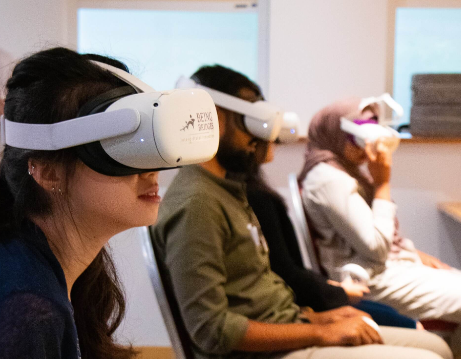 Three people sit wearing white VR headsets labeled Being Bridges in an indoor setting.