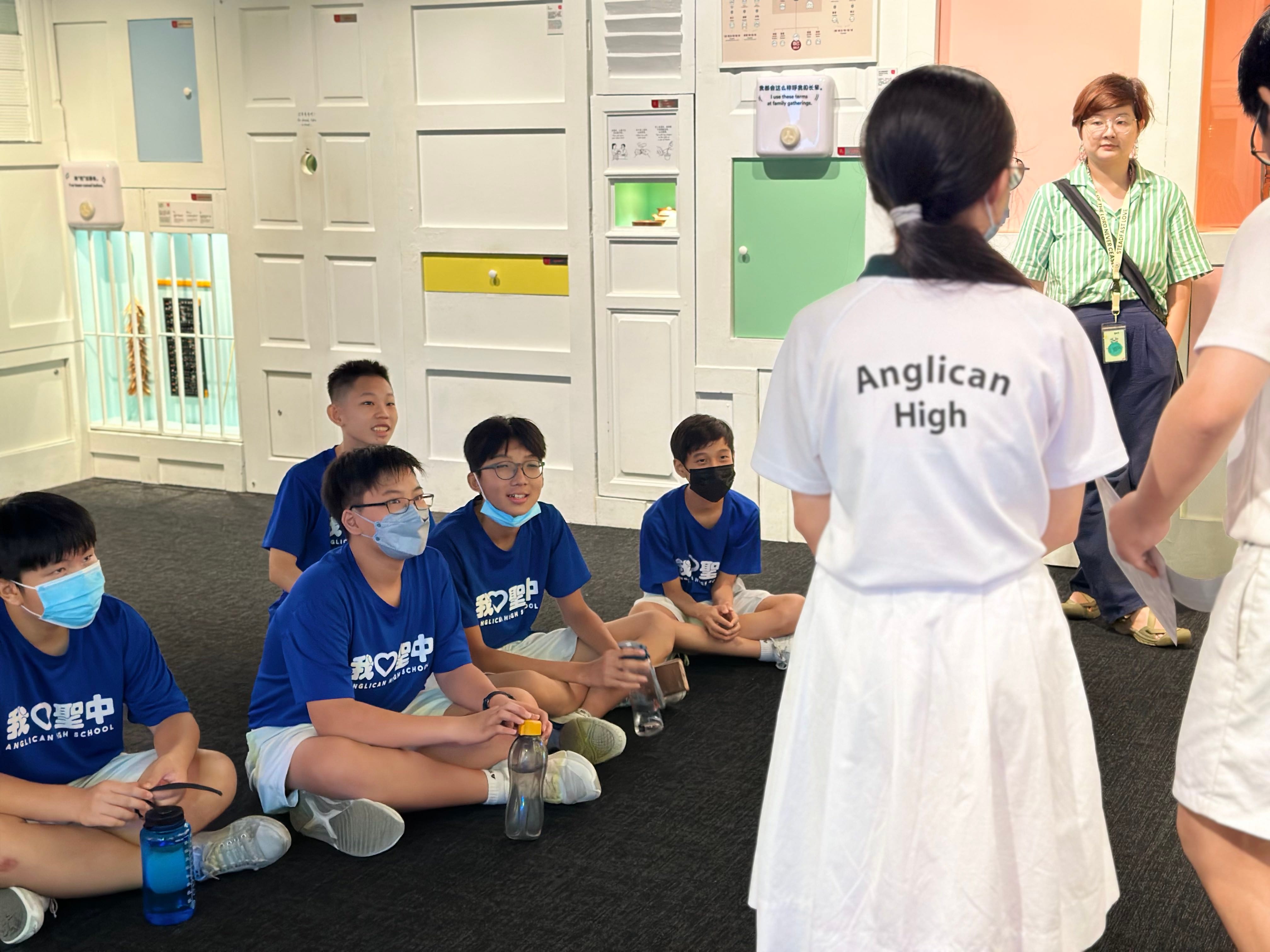 Students in blue Anglican High School shirts sit on the floor listening to a girl in a white Anglican High shirt.
