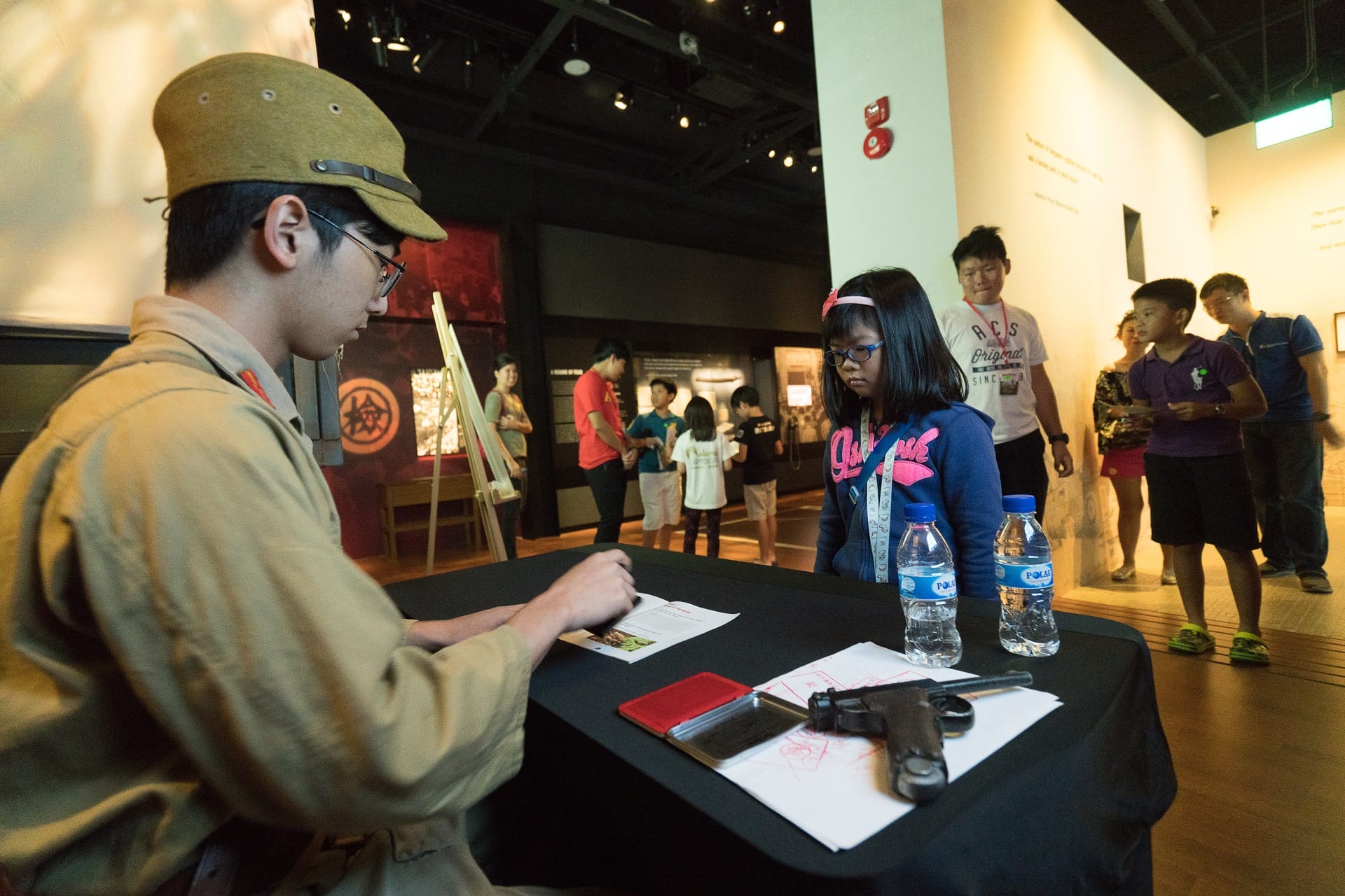 A costumed museum guide at a table with a replica pistol interacts with a girl while visitors browse exhibits behind them.