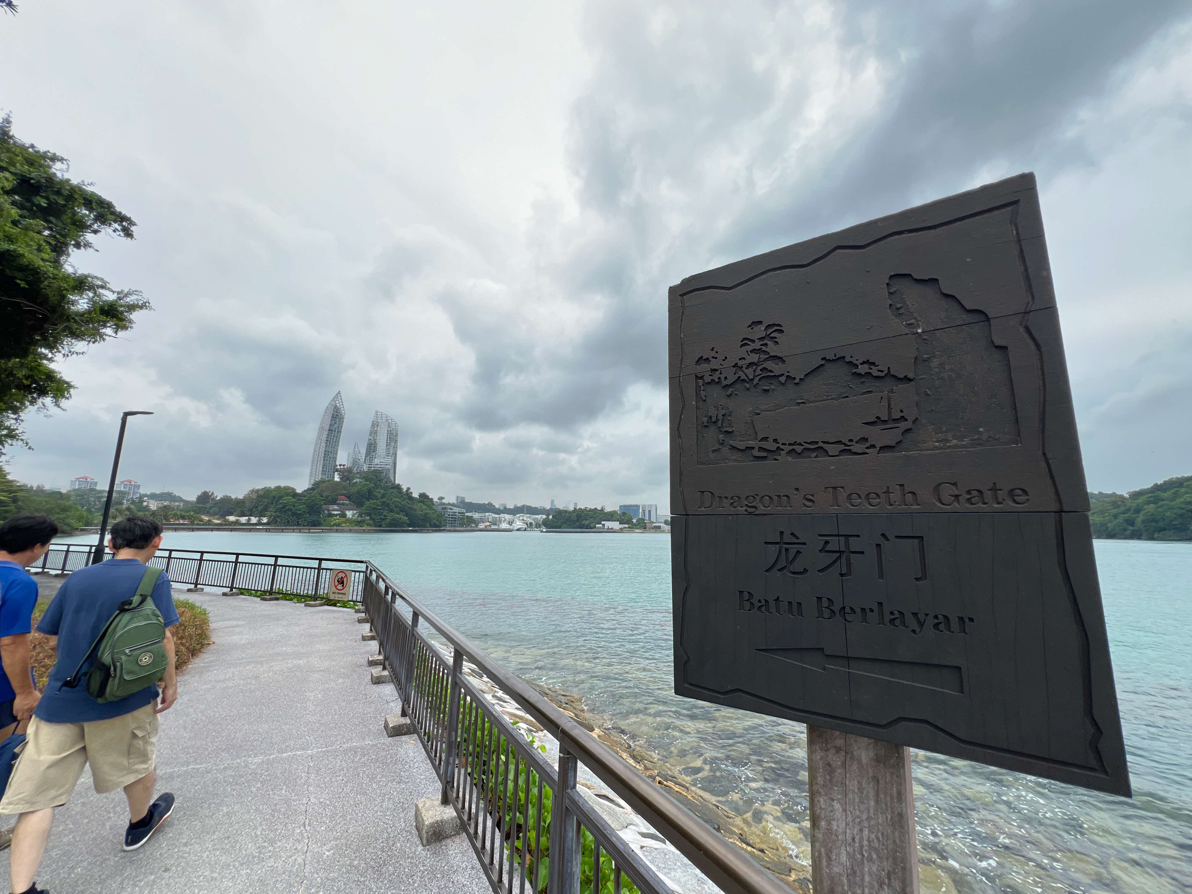 A wooden sign reading Dragon's Teeth Gate beside a waterfront path with two people walking and twin towers in the background.