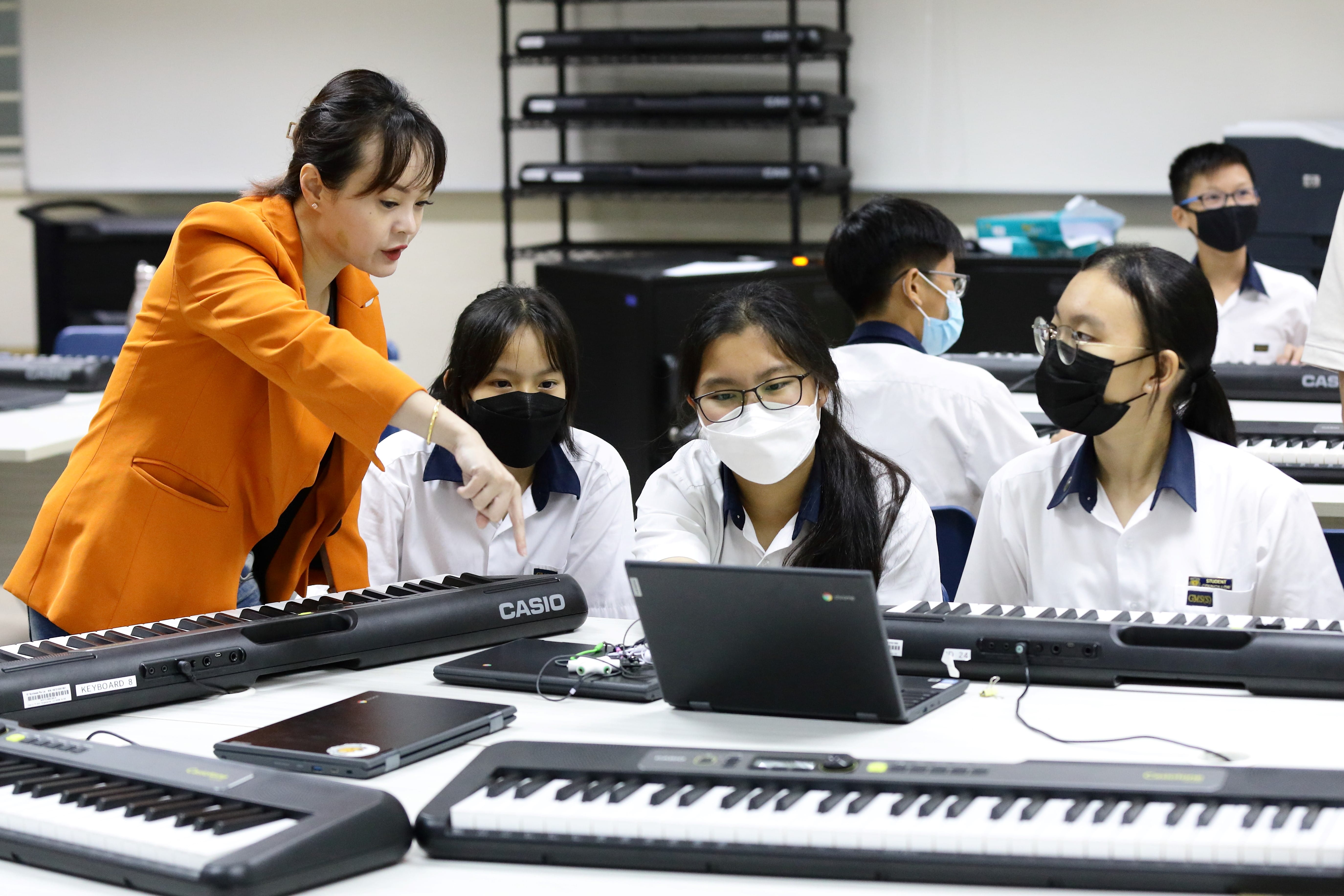 A teacher in an orange jacket points at a laptop screen while masked students sit at Casio keyboards in a music classroom.