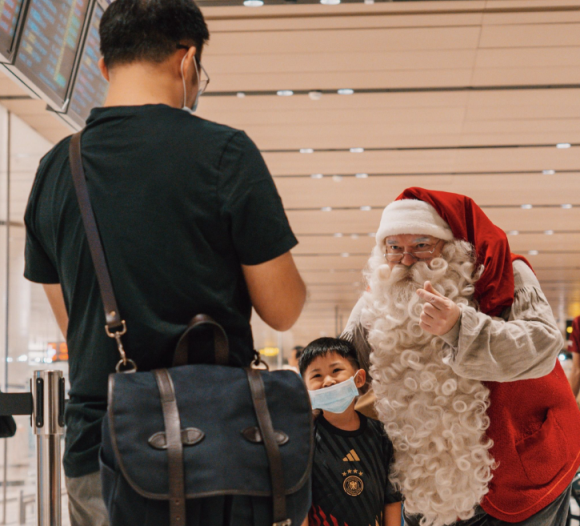 Santa Claus bends down to greet a masked child and adult at an airport terminal.