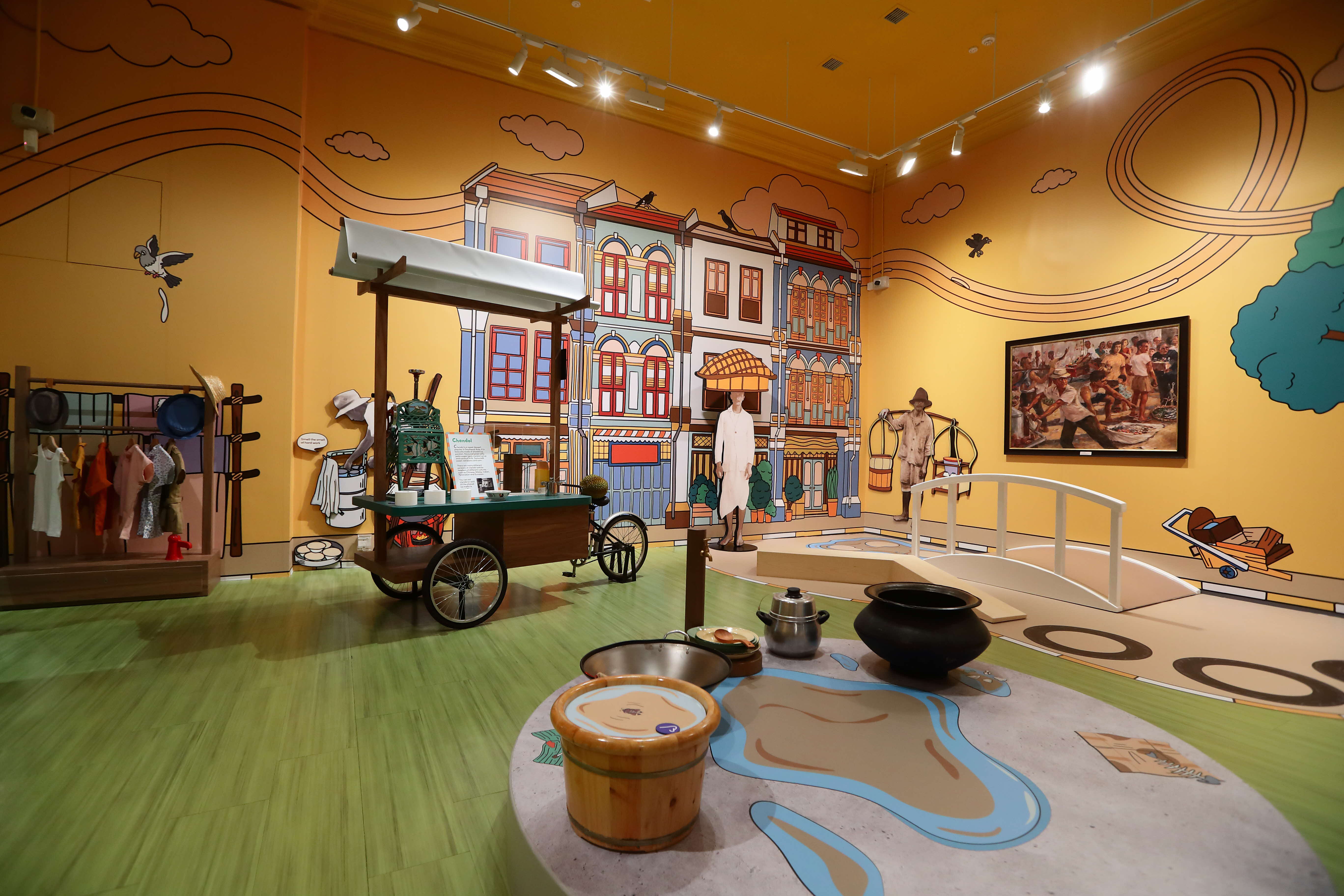 Museum exhibit with colorful cartoon street murals, a vintage food cart, mannequins, cooking pots, and clothing racks.