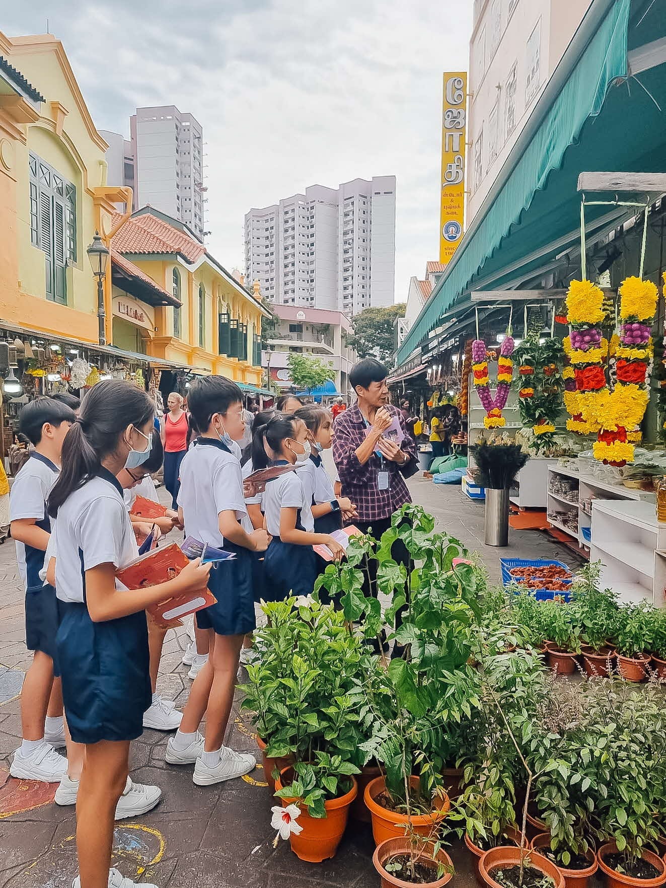Students in white and blue uniforms listen to a guide outside a flower and plant stall on a colorful urban street.