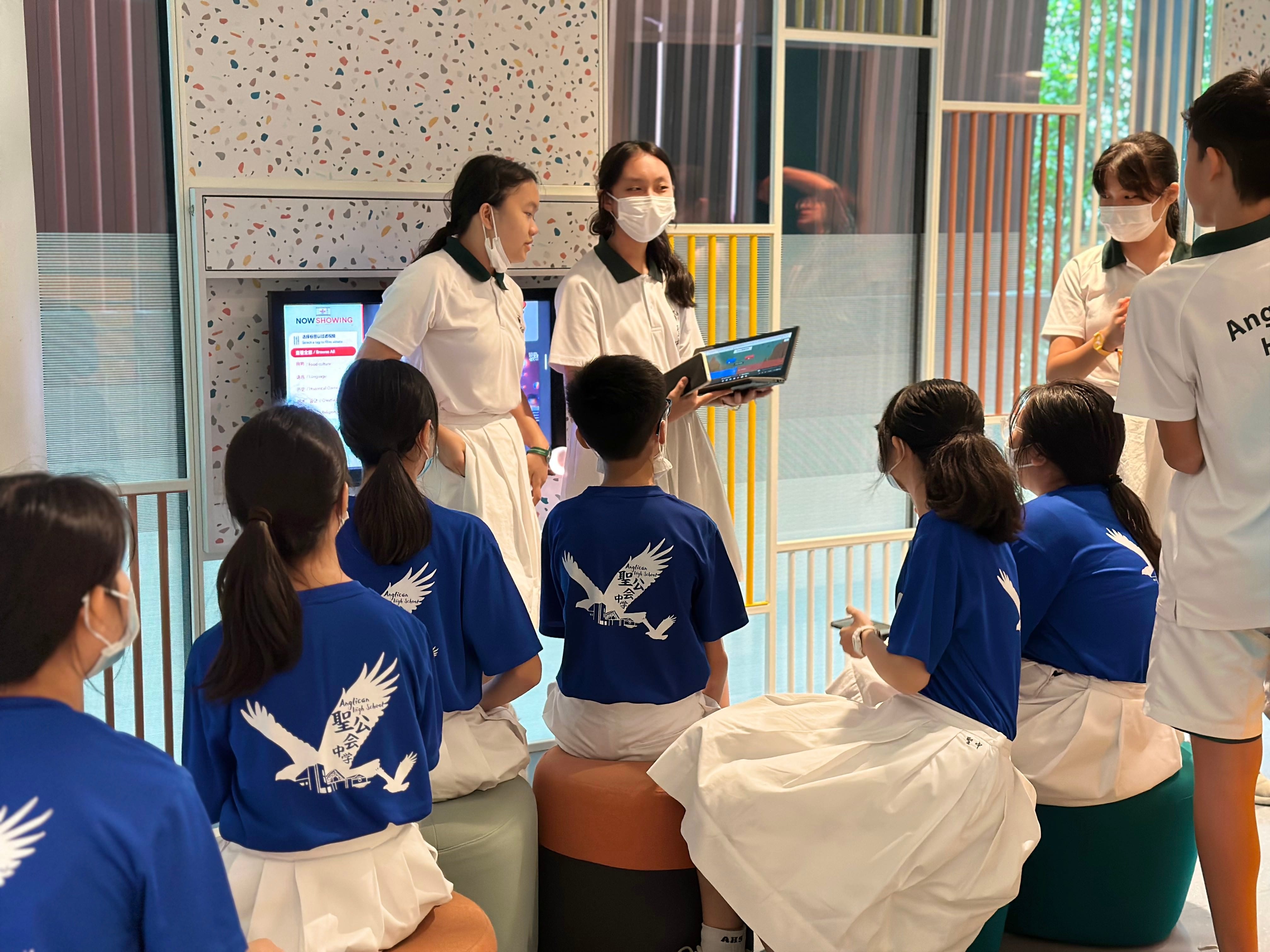 Students in blue and white uniforms gather indoors as two masked girls present using a laptop and display screen.