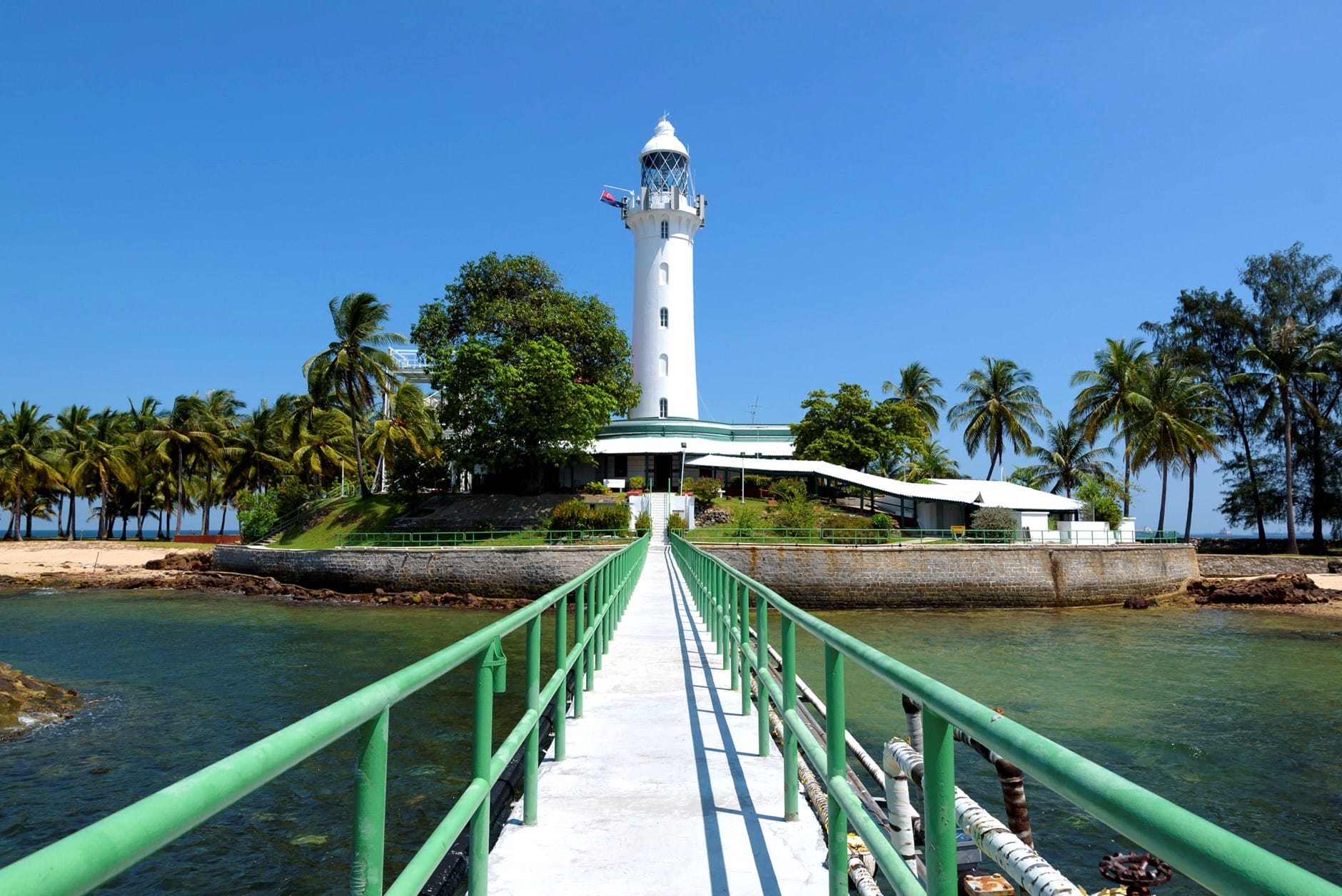 A narrow white walkway with green railings leads over water to a white lighthouse surrounded by palm trees.