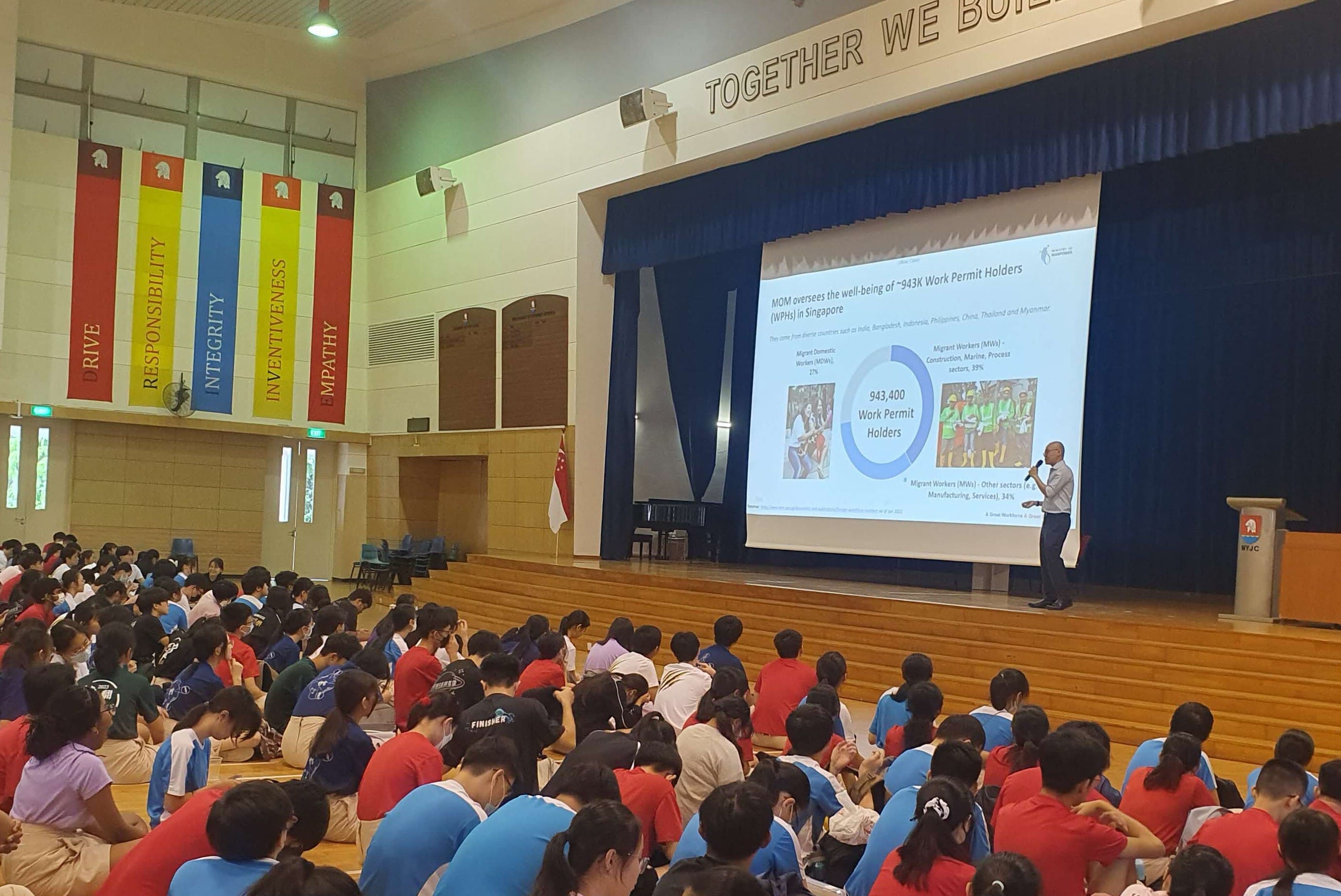 A speaker presents a slide about 943,400 Work Permit Holders in Singapore to a large audience of students in a school hall.