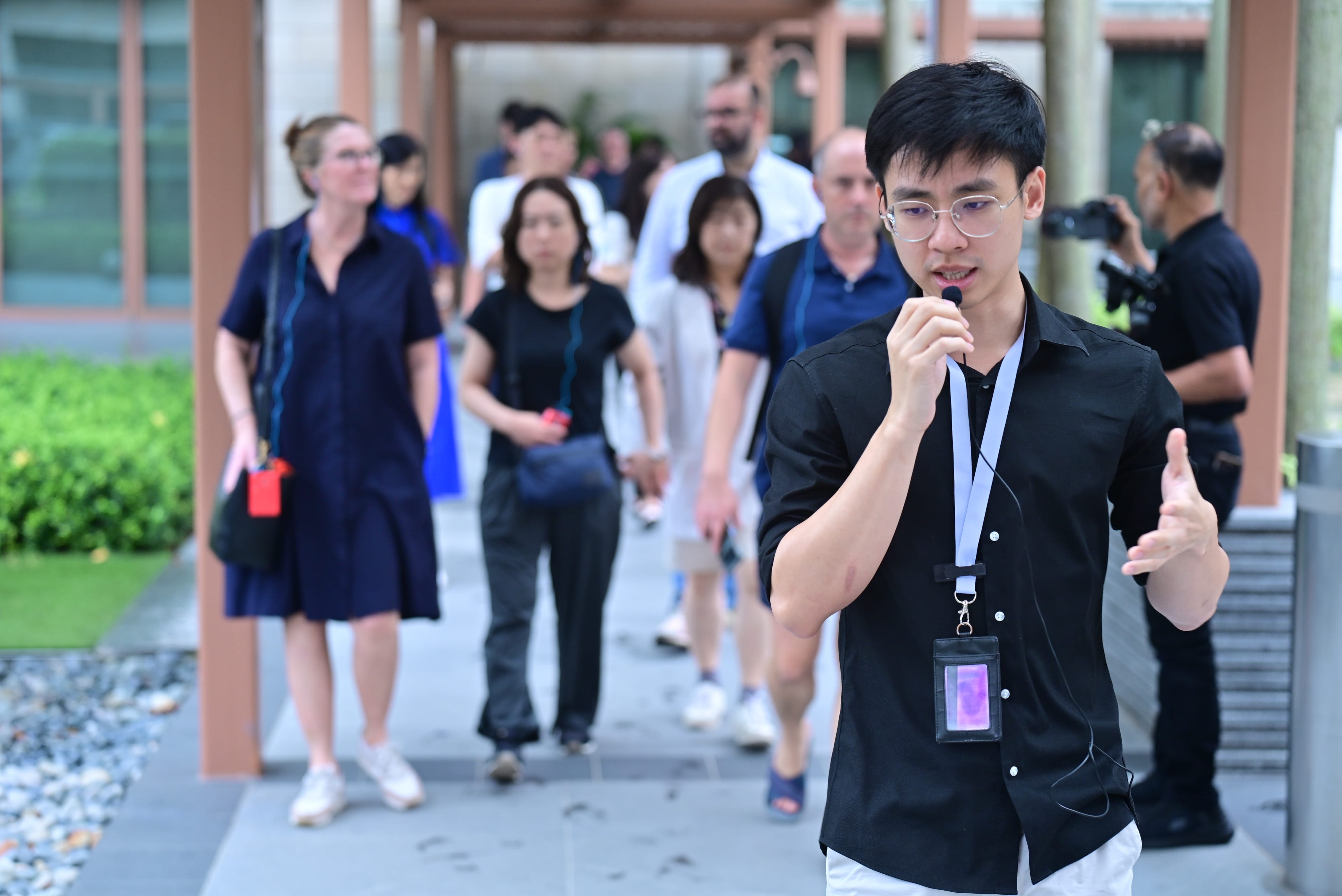 A young man with a lanyard speaks into a microphone while leading a group of people along an outdoor walkway.