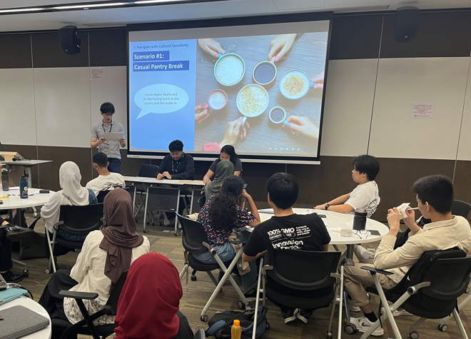 Students sit in a classroom watching a presentation slide titled "Scenario #1: Casual Pantry Break" on a large screen.