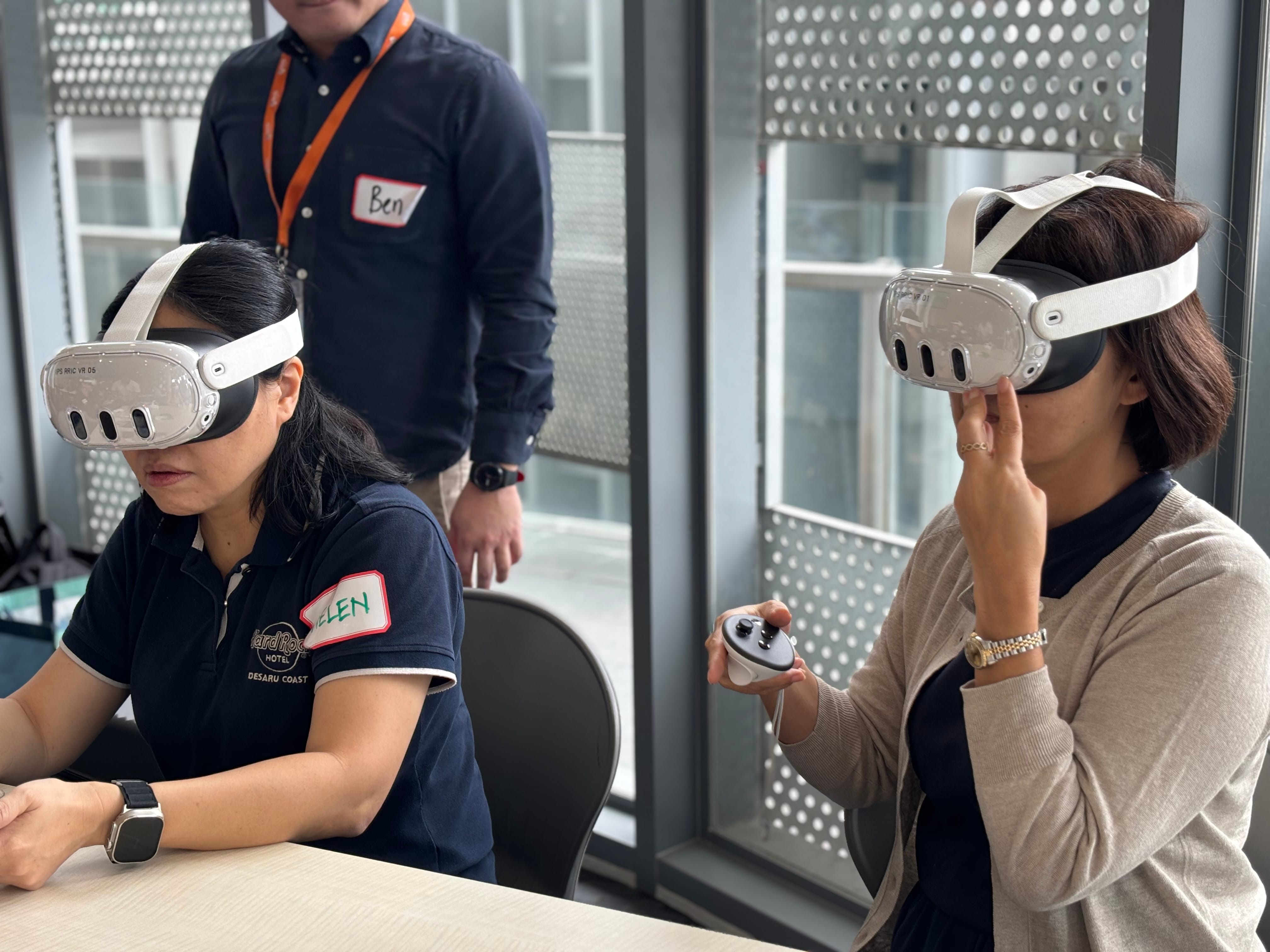 Two women wearing white VR headsets sit at a table while a man with a name tag stands behind them.