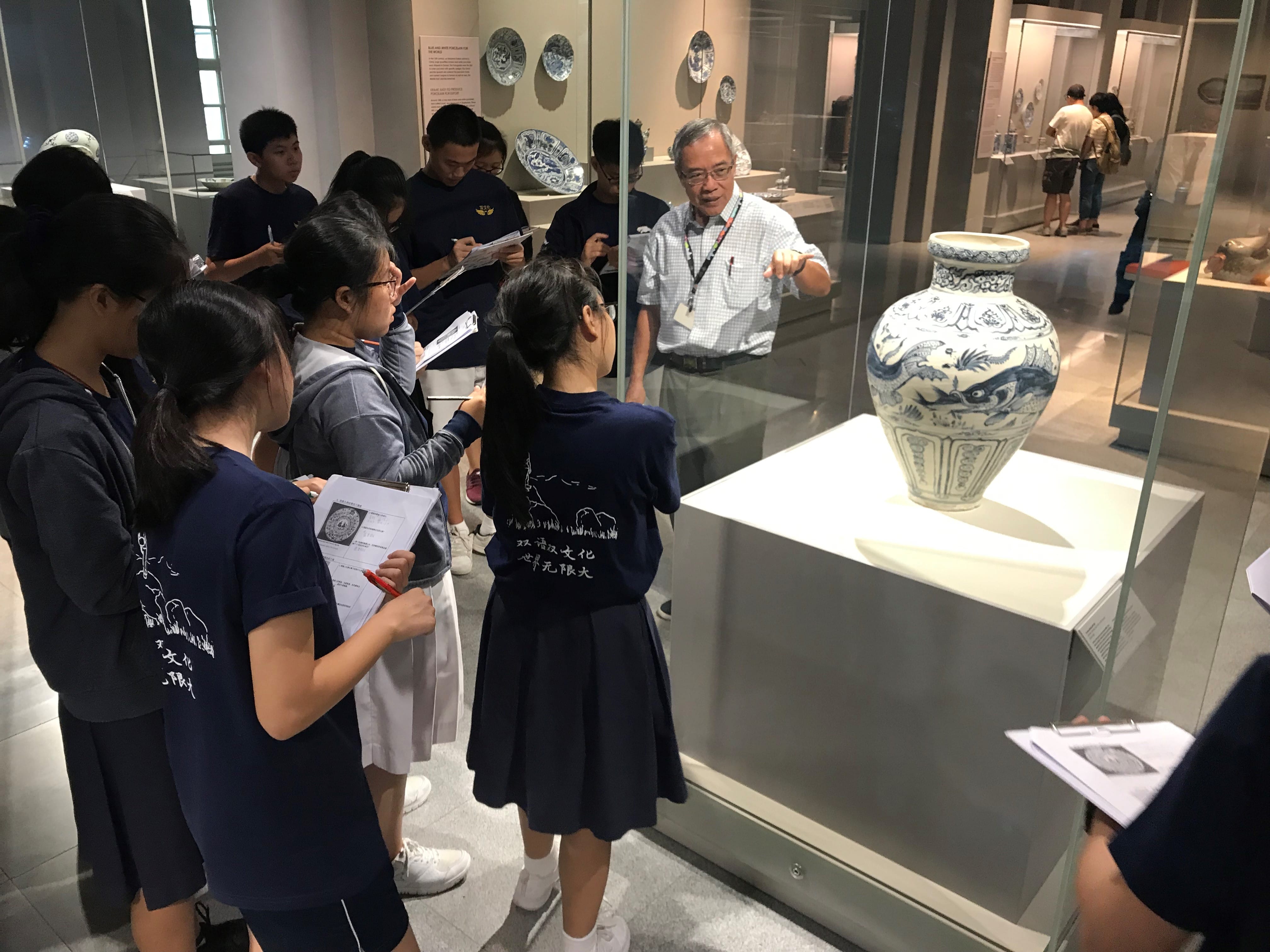 A museum guide points at a blue and white ceramic vase in a glass case while students take notes nearby.