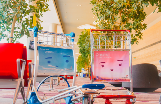 Two airport luggage carts with colorful face advertisements reading "Dreams" near indoor trees and red seating.