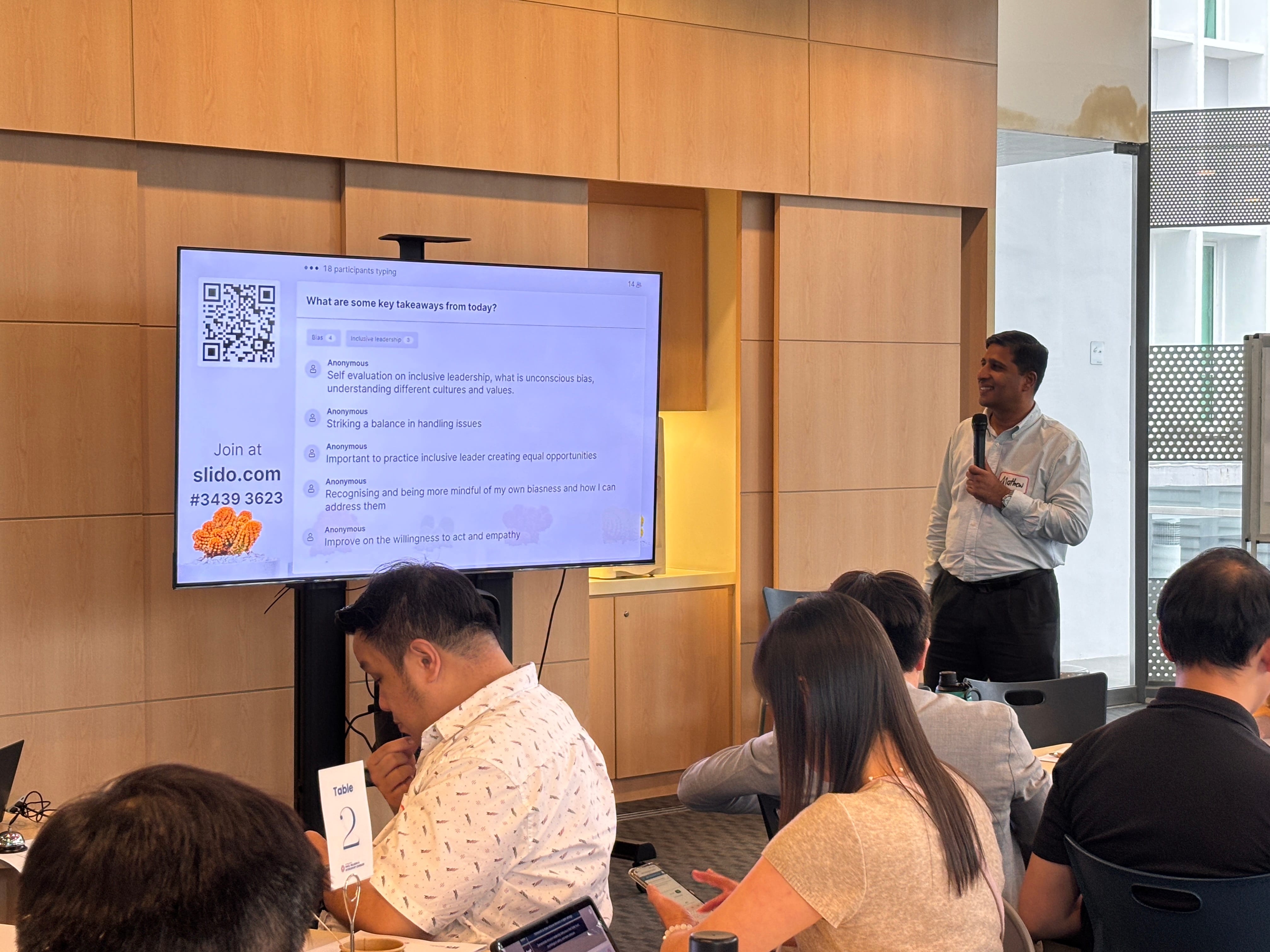A man holds a microphone presenting at a workshop, with a Slido screen showing key takeaways on inclusive leadership.