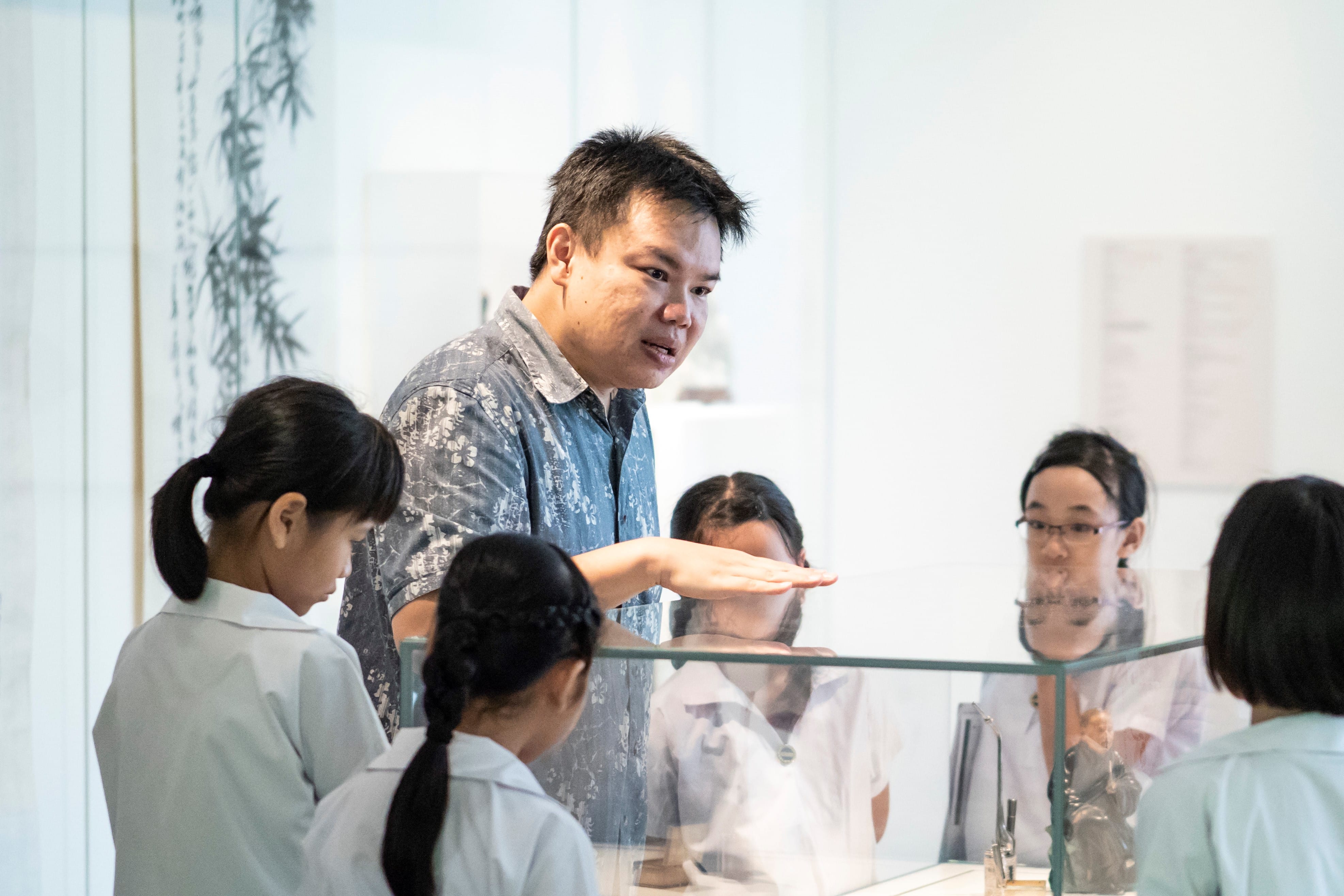 A man in a patterned shirt gestures while speaking to several people in white uniforms near a glass counter.