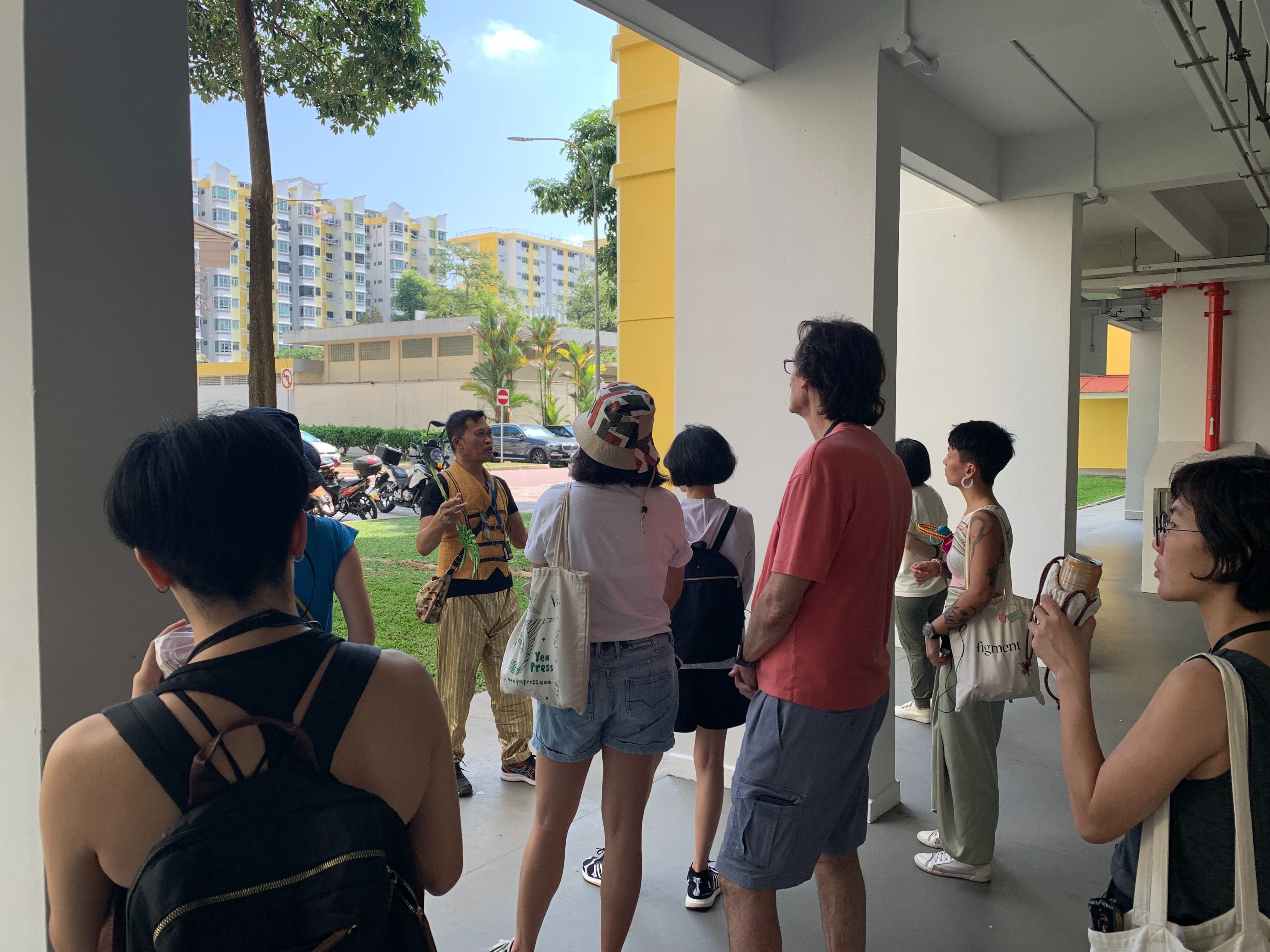 A guide in a yellow shirt speaks to a small group under a covered walkway near yellow and white apartment blocks.