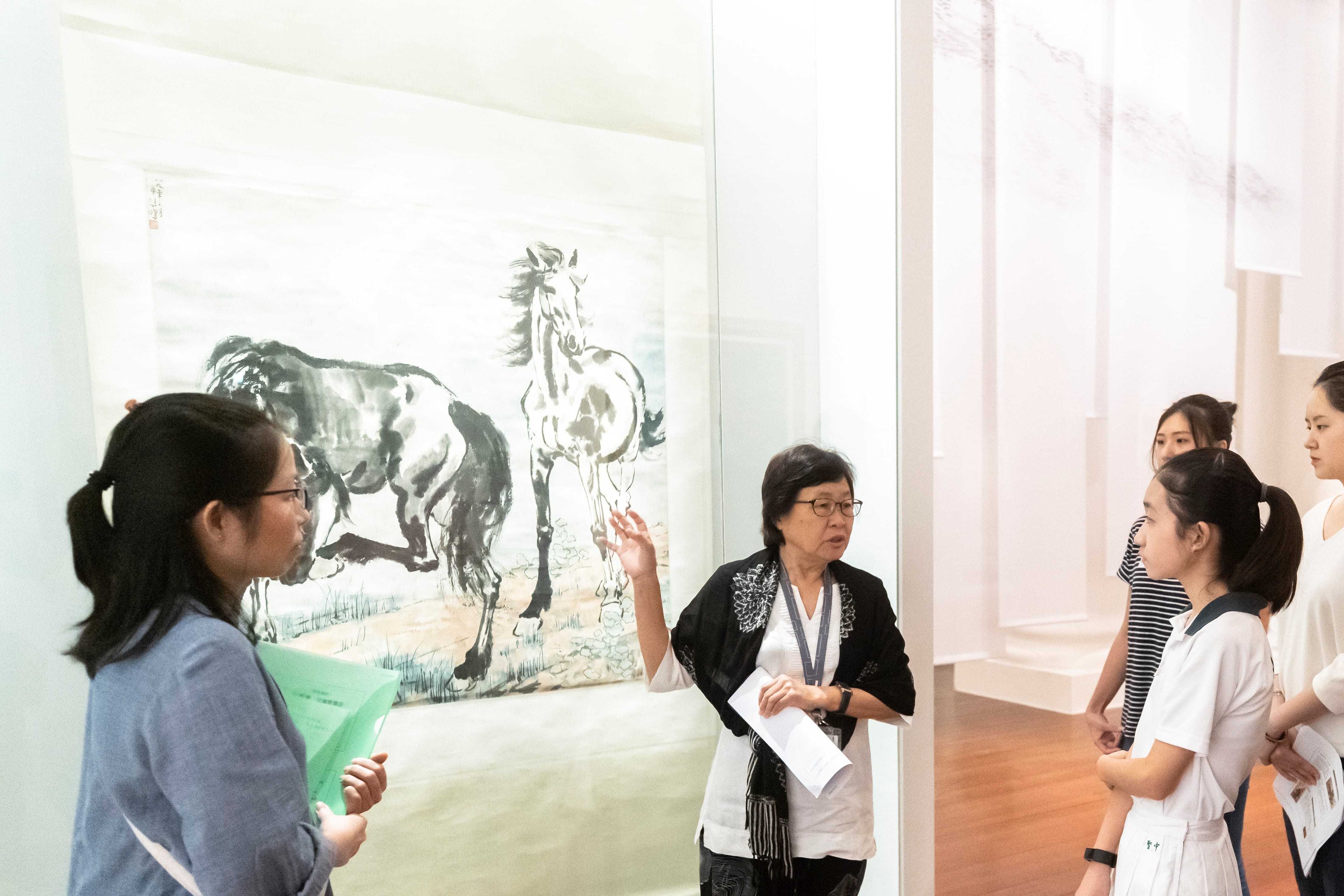 A guide speaks to a group of visitors in front of a large Chinese ink painting of two horses in a gallery.