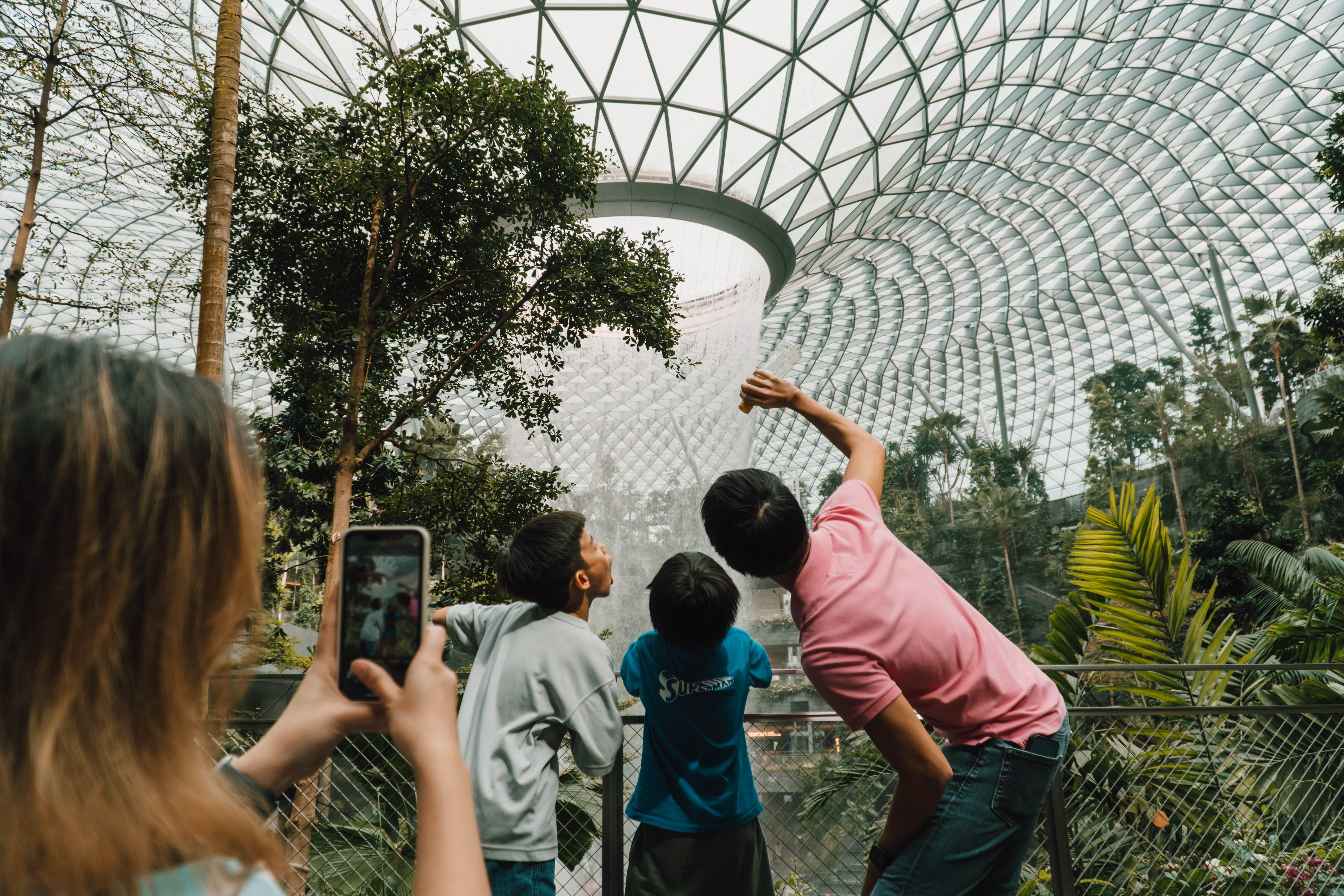 Three children look up at a large indoor waterfall under a massive glass dome structure with trees around them.