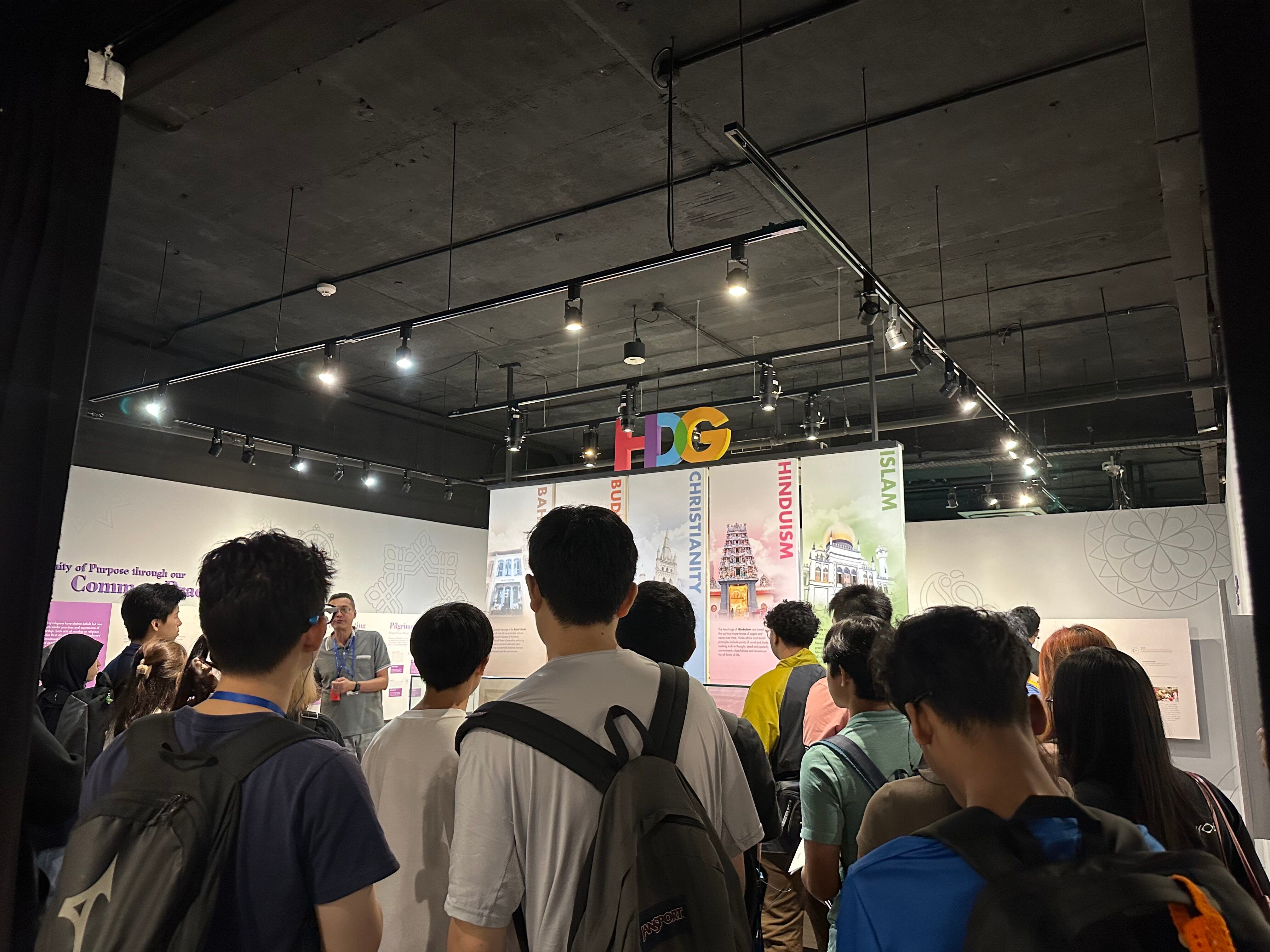 A group of visitors listens to a guide in a museum exhibit on religions including Islam, Hinduism, and Christianity.