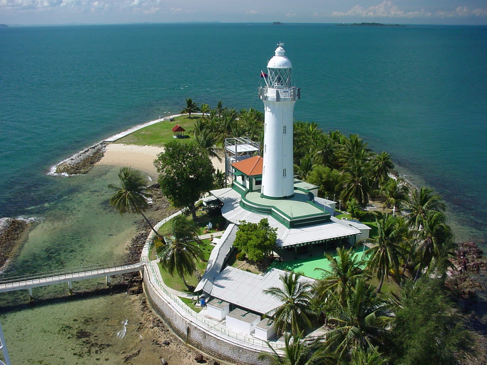 Aerial view of a white lighthouse on a small tropical island surrounded by turquoise ocean water and palm trees.