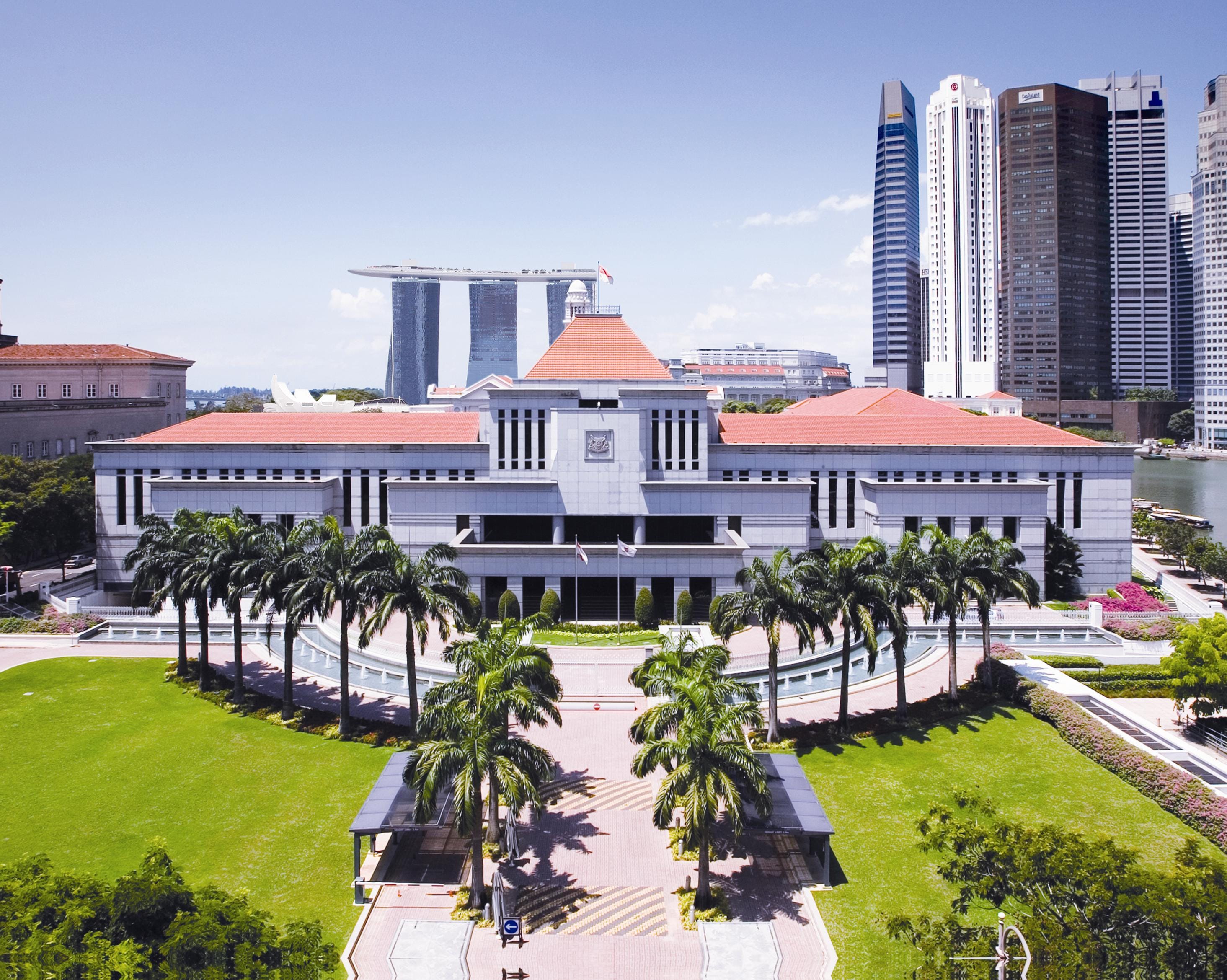 Aerial view of Singapore Parliament House with orange roof, palm trees, green lawns, and Marina Bay Sands in the background.